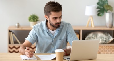 young man in blue shirt seated at table with open laptop computer, pad and paper, and cup of coffee