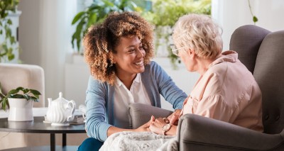 Caregiver visits with an elderly person seated in high back chair with tea pot and plants in background