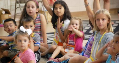 Children, ages pre-school through elementary school, seated on the floor, some with hands raised.