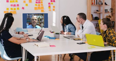 Group of people gathered around a table, watching a speaker on a screen in front of a wall full of brightly colored sticky notes.