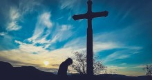 Silhouette of a person with head bowed in front of a large cross with the sun rising in the background and blue sky with wispy clouds.